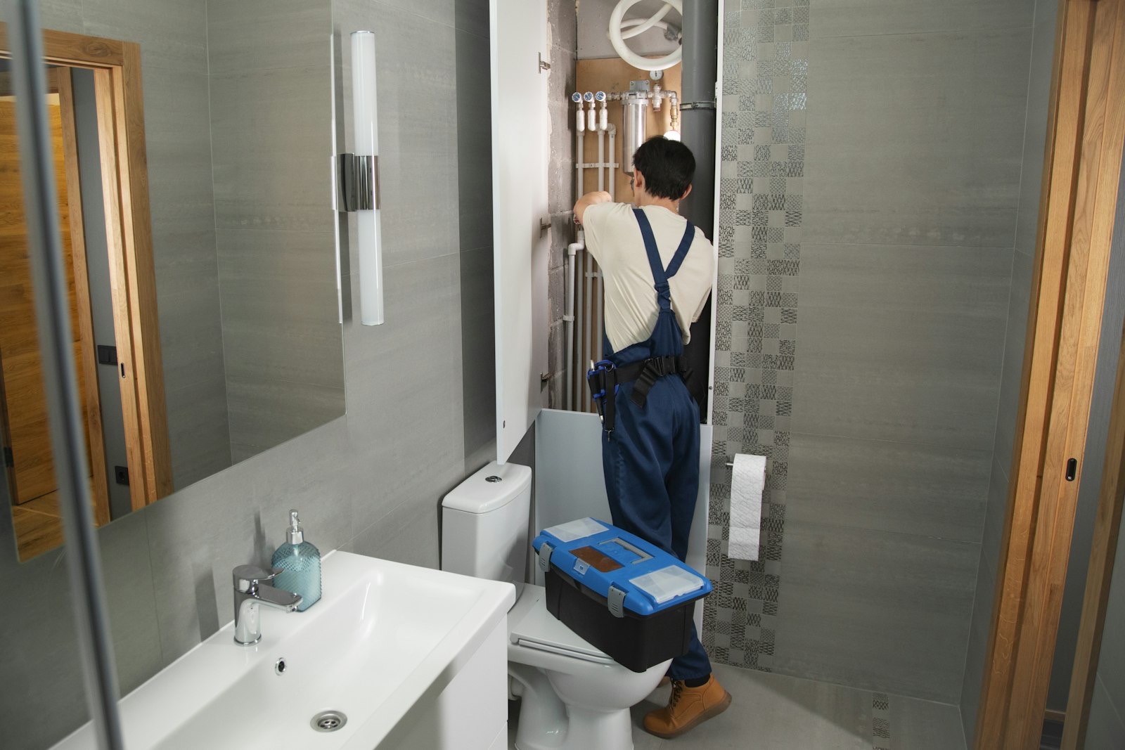 A plumber in overalls with his toolbox, working on the pipes in a bright bathroom.