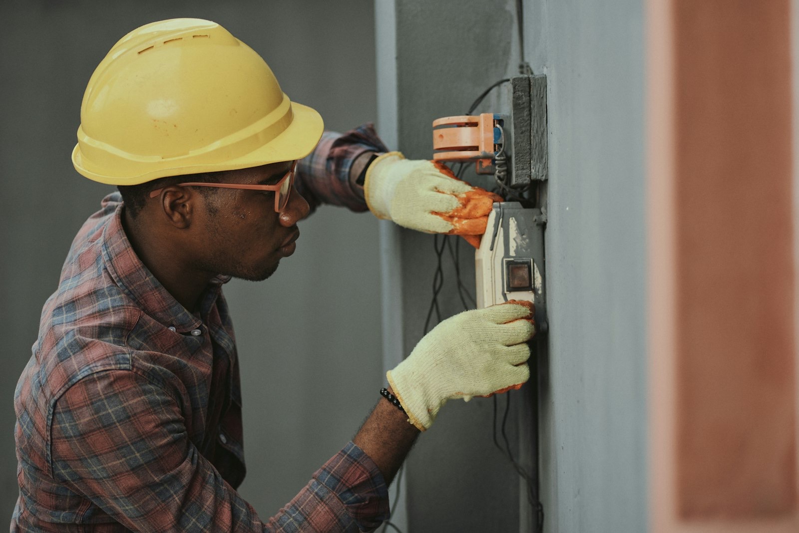 An electrician in a hard hat wiring a meter box on a wall.